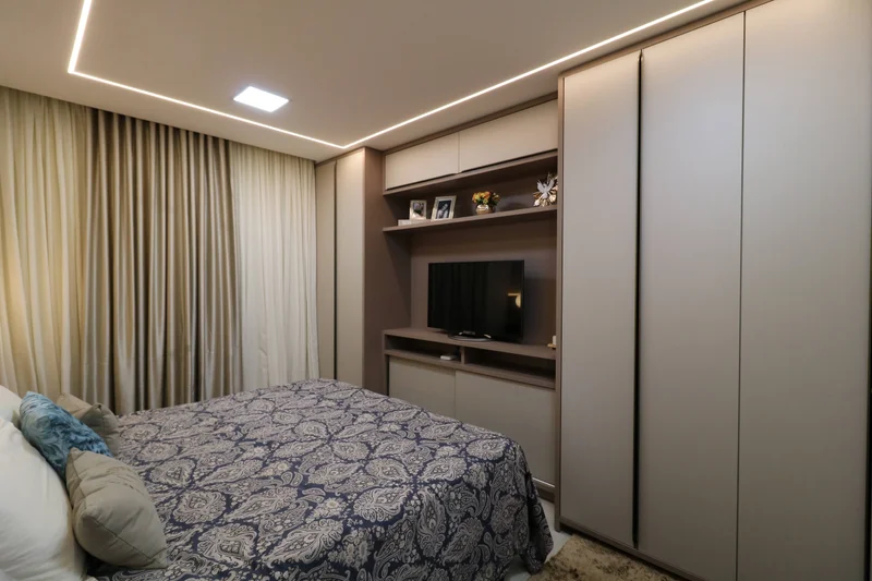Roupeiro planejado em MDF rosato e celine com portas versatile slowmotion, nicho para TV e luz indireta no quarto.